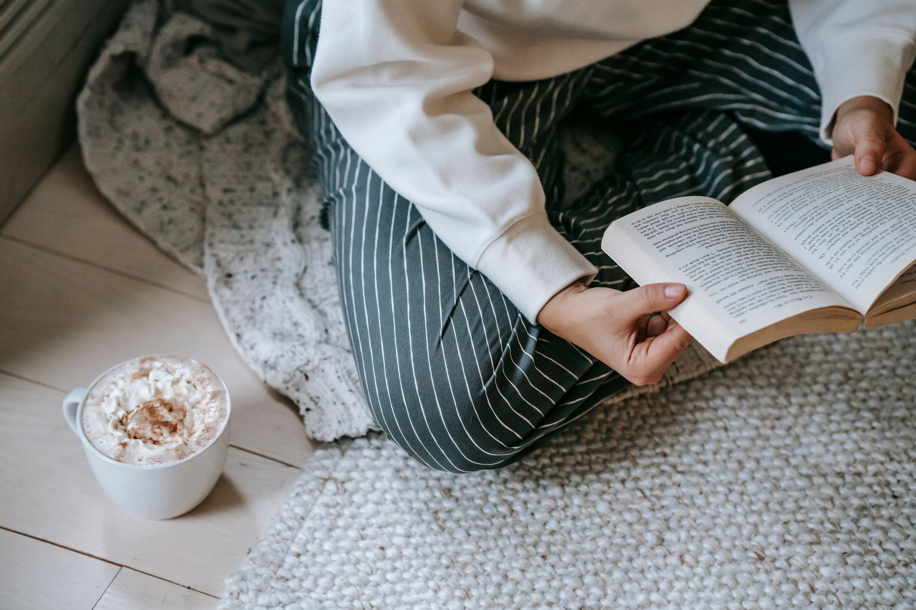 High angle of crop anonymous woman sitting on soft blanket on floor and reading literature with mug of coffee