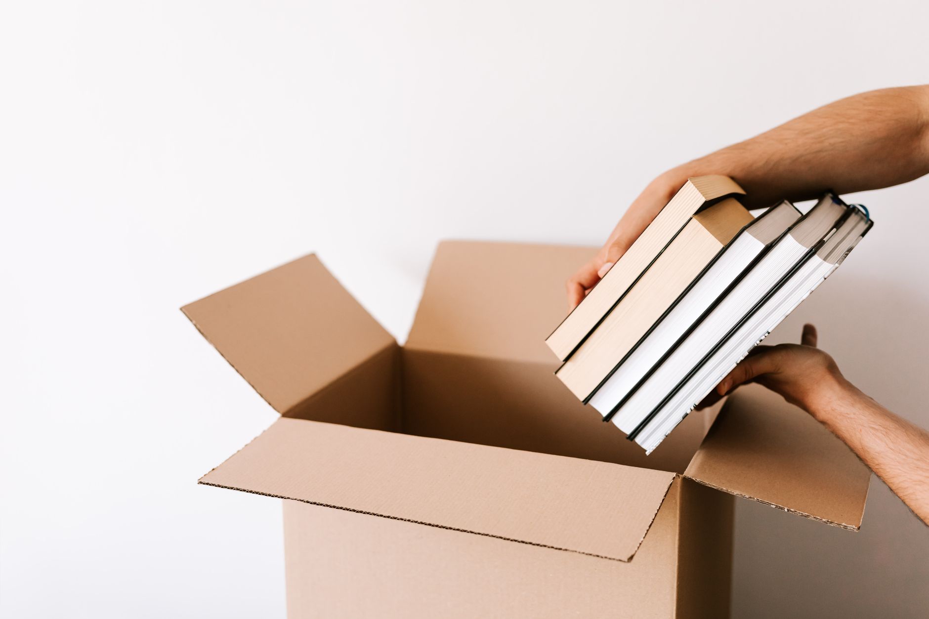 Crop unrecognizable male packing staked books into carton box while standing against white wall during relocation